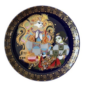 May include: A round decorative plate with a deep blue background and a gold-toned border featuring repeating circular patterns. The center displays a colorful illustration of two figures in ornate clothing, with the signature Bjorn Wiinblad visible.