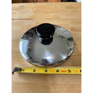 May include: A circular stainless steel pot lid with a reflective finish and a central black knob. A yellow measuring tape is positioned along the bottom edge, indicating a diameter of approximately 6 inches for the metallic cookware lid.