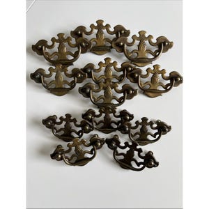 May include: A collection of nine brass drawer pulls arranged in rows. Each pull features a decorative backplate with a central pineapple motif and a curved, swinging bail handle. The metal has a darkened, aged finish with visible patina throughout.