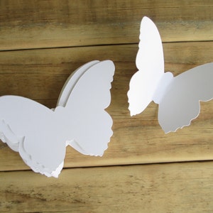 May include: White paper butterfly cutouts are displayed on a wooden surface. A stack of butterflies is on the left, and a single butterfly is on the right. The butterflies have detailed wing edges.
