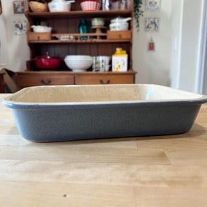 May include: A rectangular baking dish with a speckled blue exterior and a smooth, light beige interior. The dish features a wide, flat rim and deep sides, constructed from a ceramic material with a glossy finish throughout the entire piece.