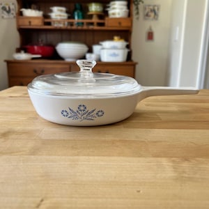以下が含まれることがあります： 長いハンドルと透明なガラスの蓋が付いた白いセラミック製のフライパン。フライパンの外側には、小さな花と葉で構成された青い花柄が中央に描かれています。蓋にはガラス製のつまみが付いています。
