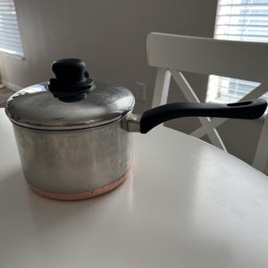 Puede incluir: Un cazo de acero inoxidable con base de color cobre, que cuenta con una tapa de acero inoxidable a juego con un pomo negro y un mango ergonómico largo y negro que se extiende desde el lateral del recipiente de cocción cilíndrico.