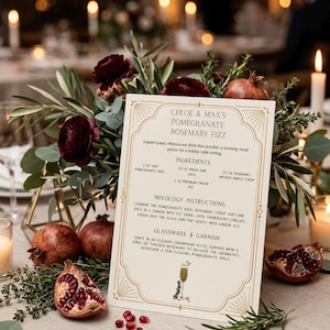 May include: A festive table setting with a cocktail recipe card for a Pomegranate Rosemary Fizz. The card is surrounded by pomegranates, burgundy flowers, and greenery. The recipe includes ingredients and instructions.