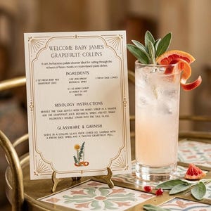 May include: A tall glass of Grapefruit Collins cocktail, garnished with sage and grapefruit peel. A recipe card titled "Welcome Baby James Grapefruit Collins" is displayed on a small gold stand. The card details ingredients and instructions for the cocktail.