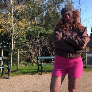 Puede incluir: Una persona lleva una chaqueta bomber marrón, una falda de punto rosa y medias negras con estampado. Lleva una gorra de béisbol marrón con un logotipo blanco. La persona está de pie frente a un banco verde y árboles.
