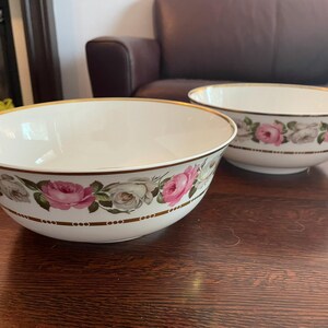 May include: Two white ceramic bowls with a gold rim and a floral rose pattern in pink and white. The bowls are on a dark wooden surface.