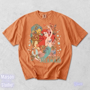 May include: Orange t-shirt featuring a colorful graphic of Ariel from The Little Mermaid, with Flounder and Sebastian. The design includes floral elements and the words "THE LITTLE MERMAID" in turquoise. The shirt has short sleeves and a crew neck.