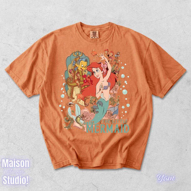 May include: Orange t-shirt featuring a colorful graphic of Ariel from The Little Mermaid, with Flounder and Sebastian. The design includes floral elements and the words "THE LITTLE MERMAID" in turquoise. The shirt has short sleeves and a crew neck.