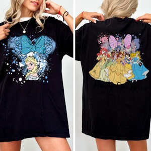 Pode incluir: Camiseta preta com um gr&aacute;fico frontal de Elsa de Frozen dentro de um design de orelha do Mickey Mouse com um la&ccedil;o azul. A parte de tr&aacute;s da camisa mostra princesas da Disney. A camisa tem um ajuste folgado.