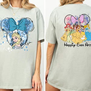 Pode incluir: Camiseta verde claro com um desenho frontal de Elsa de Frozen dentro de um contorno de orelha do Mickey, um la&ccedil;o azul e o nome "Elsa". A parte de tr&aacute;s mostra princesas da Disney com o texto "Happily Ever After".