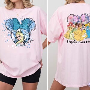 Pode incluir: Camiseta rosa claro com um design inspirado na Minnie Mouse com Elsa de Frozen, um la&ccedil;o azul e o nome "Elsa". A parte de tr&aacute;s da camiseta mostra princesas da Disney e o texto "Happily Ever After".