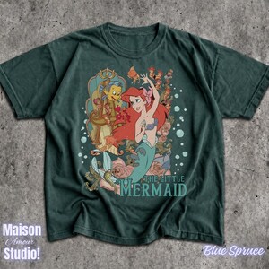 May include: A dark teal t-shirt featuring a graphic of Ariel from "The Little Mermaid." The design includes Ariel, Flounder, and other characters, with the movie title below. The shirt has the text "Maison Amour Studio!" and "Blue Spruce".