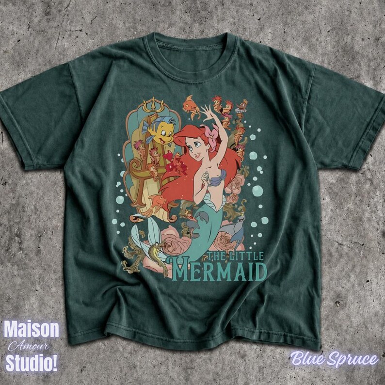 May include: A dark teal t-shirt featuring a graphic of Ariel from "The Little Mermaid." The design includes Ariel, Flounder, and other characters, with the movie title below. The shirt has the text "Maison Amour Studio!" and "Blue Spruce".