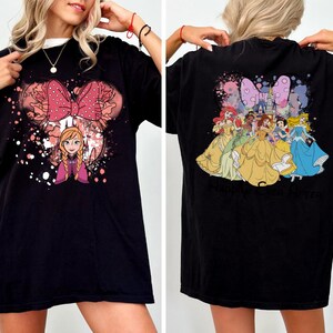 May include: Black t-shirt featuring a Minnie Mouse-inspired design with Anna from Frozen on the front. The back of the shirt displays Disney princesses. The shirt has a crew neck and short sleeves. The text "Maison Studio!" is visible on the front.