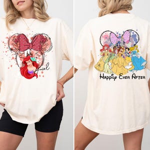 May include: Ivory-colored t-shirt featuring a Minnie Mouse-inspired design with Ariel from The Little Mermaid on the front. The back of the shirt displays Disney princesses with the text "Happily Ever After".
