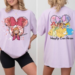May include: A light purple t-shirt featuring a Minnie Mouse-inspired design with Anna from Frozen. The front design includes a pink bow and the name "Anna". The back of the shirt displays Disney princesses and the text "Happily Ever After".