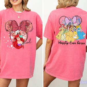 May include: A pink t-shirt featuring a Minnie Mouse design with Ariel from The Little Mermaid. The front of the shirt has Ariel's image and the name "Ariel". The back of the shirt features Disney princesses and the words "Happily Ever After".
