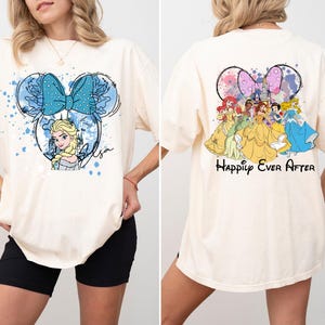 Pode incluir: Camiseta cor marfim com um design em forma de Minnie Mouse com Elsa de Frozen e um la&ccedil;o azul na frente. A parte de tr&aacute;s da camisa mostra princesas da Disney e as palavras "Happily Ever After".