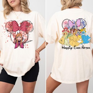 May include: Ivory-colored t-shirt featuring a Minnie Mouse-inspired design with Anna from Frozen on the front. The back of the shirt displays Disney princesses with the text "Happily Ever After". The shirt is a casual, oversized fit.