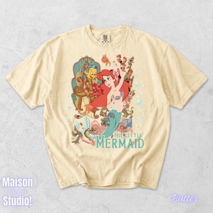 May include: A cream-colored t-shirt featuring a colorful graphic of Ariel from The Little Mermaid. The design includes Ariel, Flounder, and Sebastian, with the words "THE LITTLE MERMAID" in teal. The shirt has a vintage, slightly faded look.
