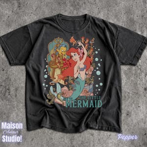 May include: Black t-shirt featuring a colorful graphic of Ariel from The Little Mermaid, with Flounder and other characters. The text "THE LITTLE MERMAID" is printed in teal. The shirt has a vintage, distressed look.