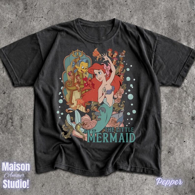May include: Black t-shirt featuring a colorful graphic of Ariel from The Little Mermaid, with Flounder and other characters. The text "THE LITTLE MERMAID" is printed in teal. The shirt has a vintage, distressed look.