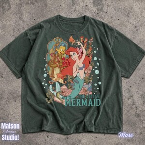 May include: Moss green t-shirt featuring a graphic of Ariel from The Little Mermaid. The design includes Ariel with red hair, Flounder, and the text "THE LITTLE MERMAID". The shirt has a vintage, distressed look.