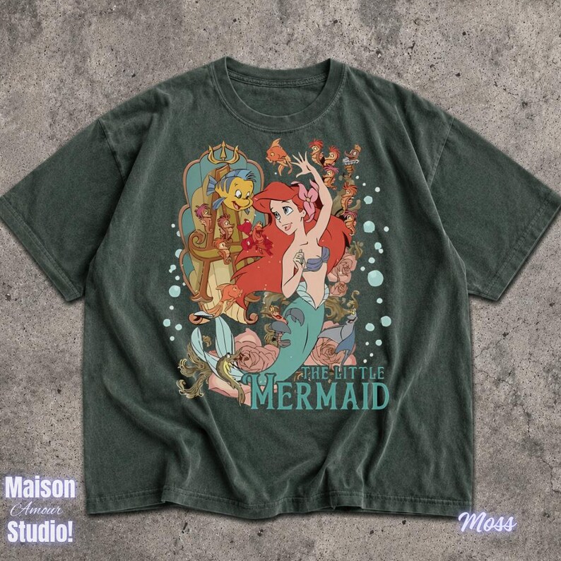 May include: Moss green t-shirt featuring a graphic of Ariel from The Little Mermaid. The design includes Ariel with red hair, Flounder, and the text "THE LITTLE MERMAID". The shirt has a vintage, distressed look.