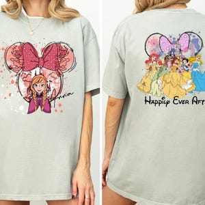 May include: A light green t-shirt featuring a Minnie Mouse-shaped design with Anna from Frozen. The front design includes a pink bow and the name "Anna". The back of the shirt displays Disney princesses with the text "Happily Ever After".