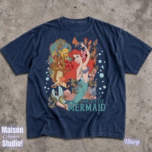 May include: Navy blue t-shirt featuring a graphic of Ariel from The Little Mermaid, with Flounder and Sebastian. The design includes the text "THE LITTLE MERMAID" in teal. The shirt has a vintage, faded look.