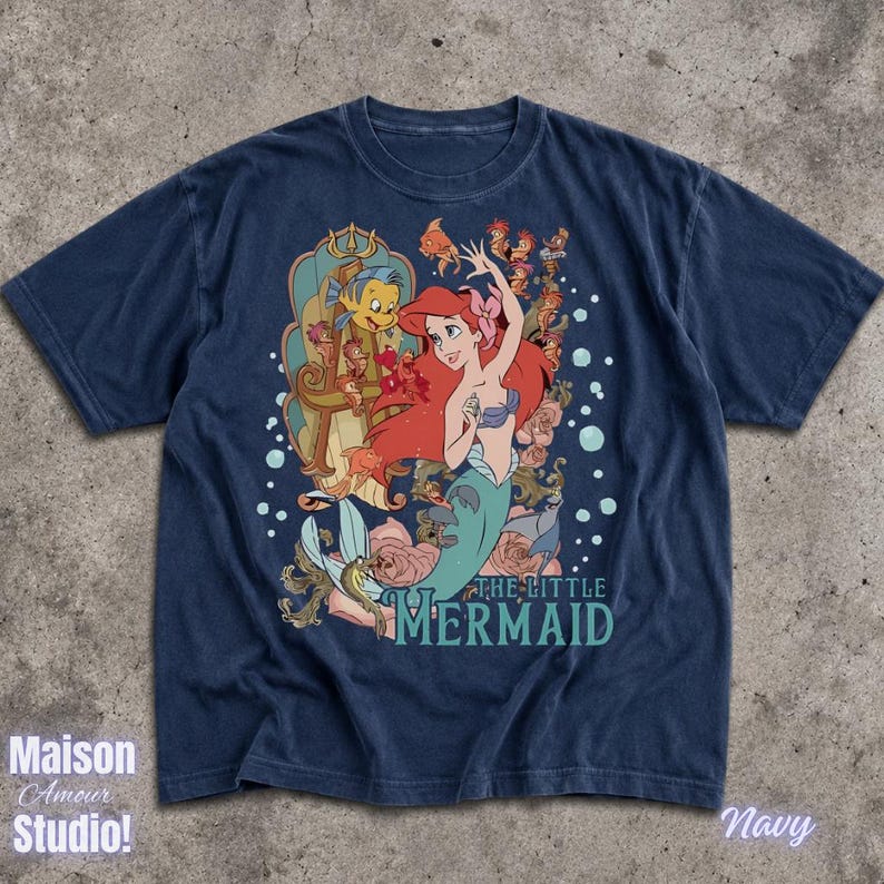 May include: Navy blue t-shirt featuring a graphic of Ariel from The Little Mermaid, with Flounder and Sebastian. The design includes the text "THE LITTLE MERMAID" in teal. The shirt has a vintage, faded look.