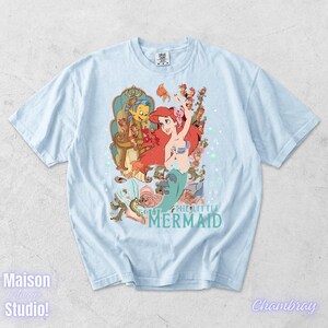 May include: Light blue t-shirt featuring a colorful graphic of Ariel from The Little Mermaid, with Flounder and Sebastian. The design includes roses and bubbles, with the words "THE LITTLE MERMAID" in a teal font. The shirt is a casual wear item.