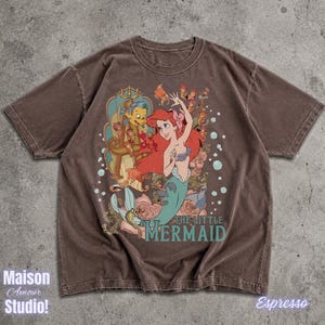 May include: Espresso-colored t-shirt featuring a graphic of Ariel from The Little Mermaid, with Flounder and other characters. The words "THE LITTLE MERMAID" are printed below the image. The shirt also has the text "Maison Amour Studio!"
