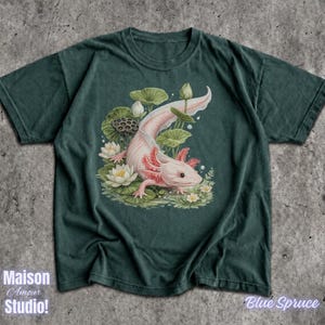 Könnte beinhalten: Dunkelblaugrünes T-Shirt mit einer Aquarellillustration eines rosa Axolotls inmitten von Seerosen und Blumen. Der Text "Maison Amour Studio!" befindet sich links und "Blue Spruce" rechts. Der Axolotl hat rote Kiemen.