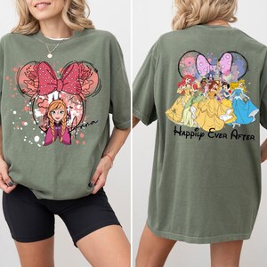 May include: Olive green t-shirt featuring a Minnie Mouse-inspired design with Anna from Frozen on the front. The back of the shirt displays Disney princesses and the text "Happily Ever After". The shirt has a relaxed fit.