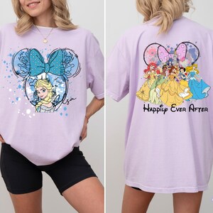 Pode incluir: Uma camiseta lil&aacute;s clara com um design inspirado na Minnie Mouse, apresentando Elsa de Frozen, um la&ccedil;o azul e o nome "Elsa". A parte de tr&aacute;s da camiseta mostra princesas da Disney e o texto "Happily Ever After".