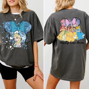 Pode incluir: Camiseta cinza escuro com um design da Minnie Mouse com Elsa de Frozen e um la&ccedil;o azul na frente. A parte de tr&aacute;s da camisa mostra princesas da Disney com o texto "Happily Ever After".