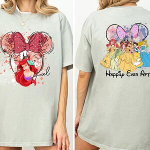 May include: A light green t-shirt featuring a Minnie Mouse-shaped design with Ariel, a red bow, and the name "Ariel". The back of the shirt displays Disney princesses and the words "Happily Ever After".