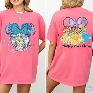 Pode incluir: T-shirt rosa com um desenho animado de Elsa e um la&ccedil;o azul. A parte de tr&aacute;s da camisa apresenta princesas da Disney e as palavras "Happily Ever After".