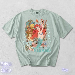 May include: Light green t-shirt featuring a colorful graphic of Ariel from The Little Mermaid. The design includes Ariel, Flounder, and Sebastian, with the words "The Little Mermaid" in a teal font. The shirt has short sleeves and a crew neck.