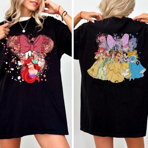 May include: Black t-shirt featuring a Minnie Mouse design with a red bow and Ariel the Little Mermaid. The back of the shirt displays several Disney princesses. The shirt has the text "Maison Studio!" on the front and "Black" on the back.
