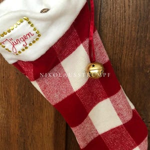 Op de afbeelding: Rode en witte kerstsok met buffalo plaid-patroon, een witte boord en gouden details. De sok heeft een gouden bel aan de bovenkant en de naam "Jungen" geborduurd op de boord.