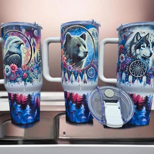 May include: Three white stainless steel travel mugs with colorful designs. The mugs feature a wolf, a bear, and an eagle with dreamcatchers and feathers. The mugs have lids and handles.