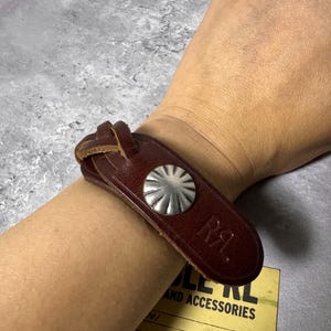 May include: A dark brown leather bracelet featuring a braided strap design, a silver-toned circular metal concho button, and the embossed letters RRL. The leather band is secured around the wrist, showcasing a smooth finish and visible stitching.