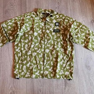 May include: A long-sleeve jacket featuring an olive green and white camouflage pattern. The garment has a full-length front zipper, a collar, and two black rectangular patches on the chest, one displaying the letter H and the other the text APE.