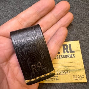 May include: A dark brown leather money clip featuring a stamped sunburst pattern and the letters RRL embossed on the front. The clip is secured with thick, light-colored stitching at the bottom edge and is displayed alongside a tan paper tag.