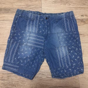 May include: Blue denim shorts featuring a patchwork design with various patterns including anchors, paisley, and stripes. The shorts have a button closure, a zip fly, belt loops, and a faded wash finish across the different fabric panels.