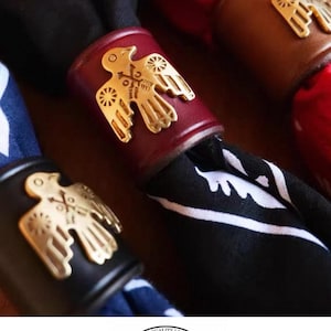 Op de afbeelding: Drie lederen servetringen in zwart, bordeaux en bruin, elk voorzien van een goudkleurig metalen vogel-embleem. De ringen houden gevouwen stoffen servetten vast. Onder de items staat een cirkelvormig logo met de tekst: FINEST QUALITY LEATHERS HORWEEN.