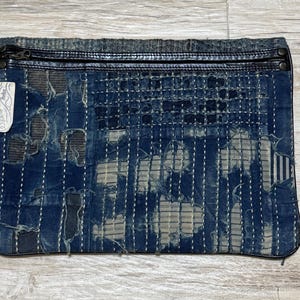 Può includere: Una pochette rettangolare con cerniera realizzata in tessuto tinto indaco usurato, caratterizzata da visibili cuciture bianche in stile sashiko, parti sfilacciate e un bordo in pelle scura lungo il lato superiore. È presente una piccola etichetta.